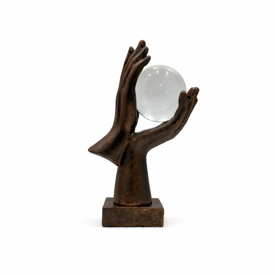 Bronze-effect hands holding a clear crystal sphere on a rustic wooden side table next to a stack of books.