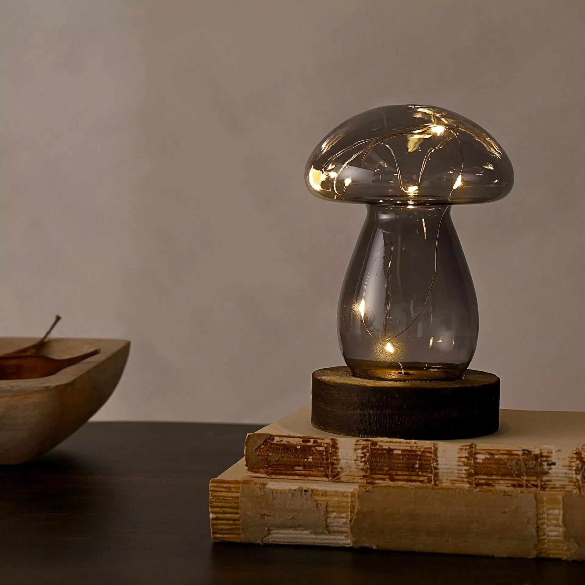 Grey Glass Mushroom on a oak wood side table on top of 2 books with a taupe wall background